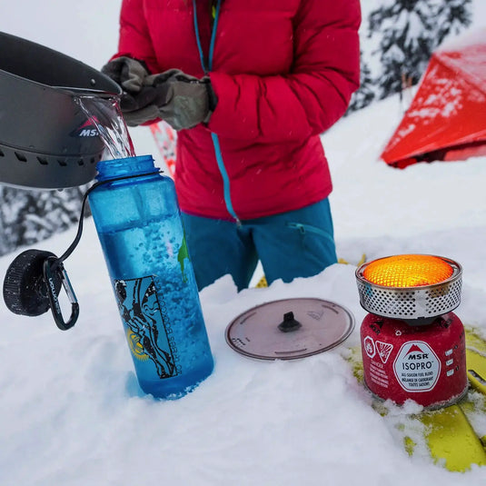 Person in roter Jacke, die ein MSR® Reactor® Kochsystem, bekannt als der effizienteste tragbare Kocher, verwendet, um Schnee zu Wasser zu schmelzen, mit einem Brennstoffkanister und einer Wasserflasche auf dem Schnee