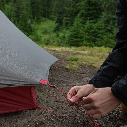 Eine Person sichert die Abspannleine eines MSR® FreeLite™ 3 „V3“ auf einem bewaldeten Campingplatz.