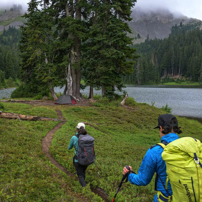 Zwei Wanderer nähern sich mit ihrem ultraleichten Zelt MSR® FreeLite™ 3 „V3“ einem Campingplatz an einem See, umgeben von Wald und nebelbedeckten Bergen.