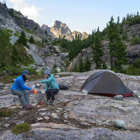 Zwei Camper bereiten eine Mahlzeit in der Nähe eines MSR® FreeLite™ 3 „V3“ vor einer bergigen Kulisse in der Abenddämmerung zu.