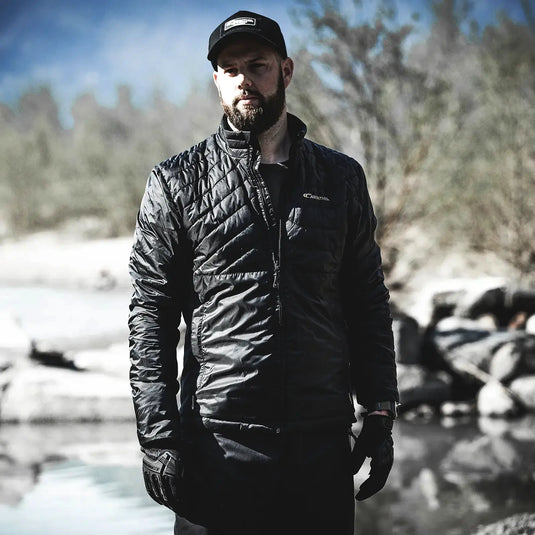 Mann in schwarzer Carinthia® G-LOFT® Ultra Jacket 2.0 und Mütze steht draußen in der Nähe eines Gewässers.