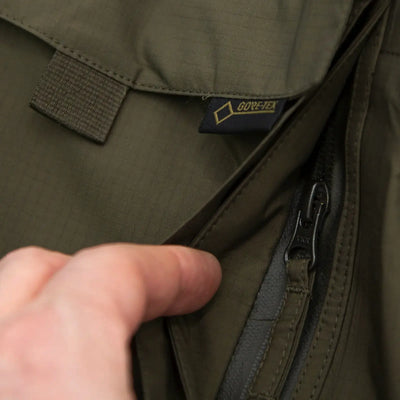 Eine Hand öffnet einen Reißverschluss an einer atmungsaktiven, grünen Carinthia® TRG Jacket mit sichtbarem 'Gore-Tex'-Etikett.