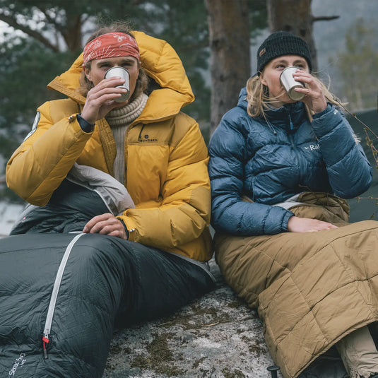 Zwei Personen in warmer Robens® Schlafsack Serac 600 -14°C Kleidung sitzen draußen und trinken aus Tassen.