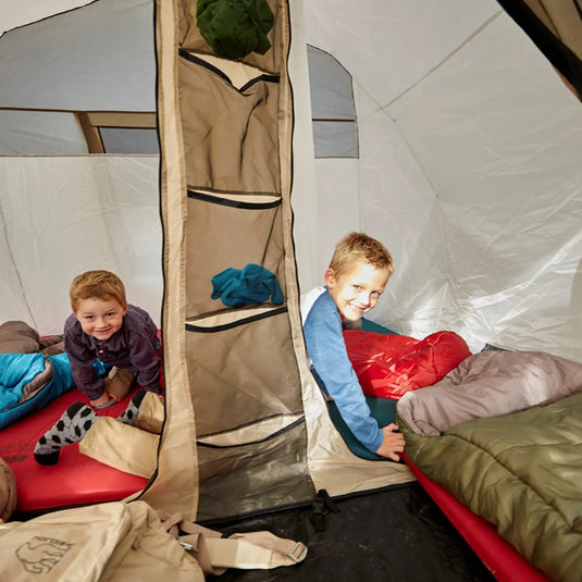 Zwei lächelnde Kinder in einem hellen Campingzelt mit Schlafsäcken und einer selbstaufblasenden Matratze vom Typ Grand Canyon® Hattan 3.8.