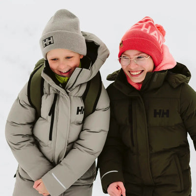 Zwei Junioren in warmen Adore Puffy Parkas von Helly Hansen® Juniors lächeln und genießen die gemeinsame Zeit im Freien.