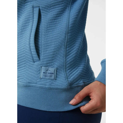 Nahaufnahme eines blau strukturierten Lillo Outdoor-Pullovers für Damen von Helly Hansen® mit einem kleinen Helly Hansen-Logoaufnäher und einer Person, die den Saum festhält.