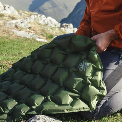 Person in einer orangefarbenen Jacke bläst im Freien eine grüne XL HIGHLANDER® NAP-PAK aufblasbare Isomatte auf.