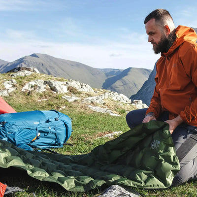 Mann baut einen Campingplatz in den Bergen HIGHLANDER NAP-PAK Aufblasbare Isomatte, XL auf.