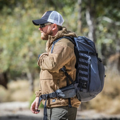 Ein Mann mit Mütze, Sonnenbrille, Helikon-Tex® Cumulus Hochleistungsfleecejacke und Rucksack steht in einem Waldgebiet.