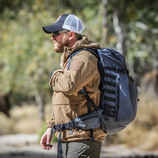 Ein Mann mit Mütze, Sonnenbrille, Helikon-Tex® Cumulus Hochleistungsfleecejacke und Rucksack steht in einem Waldgebiet.