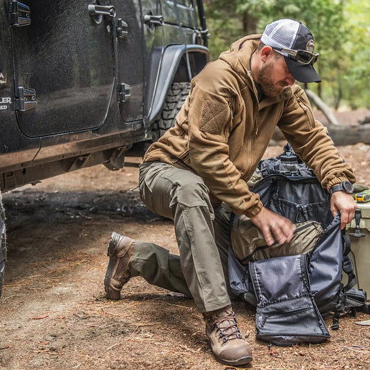 Ein Mann kauert neben einem Fahrzeug im Wald und packt Ausrüstung aus einem Helikon-Tex® Cumulus Hochleistungsfleecejacke-Rucksack ein oder aus.