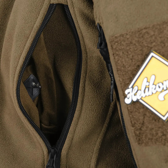 Nahaufnahme einer grünen Helikon-Tex® Liberty Jacke – Double Fleece mit teilweise geöffnetem schwarzen Reißverschluss und einem „Helikon-Tex“-Markenaufnäher am Ärmel.