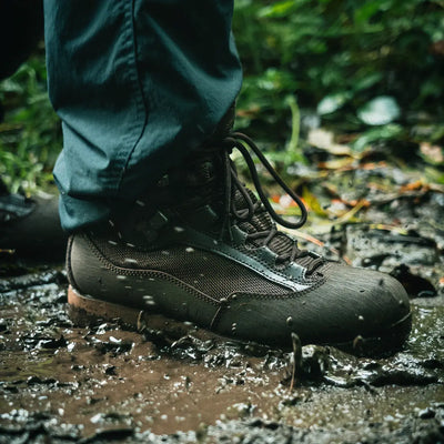 Person mit wasserdichten HIGHLANDER® „Pentland“ Stiefeln tritt auf einem nassen Waldweg in eine Pfütze.