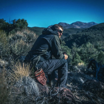 Ein Wanderer mit Sonnenbrille und Helikon-Tex® Woodsman Anorak Jacket® macht eine Pause an einem felsigen Hang mit Bergen im Hintergrund.