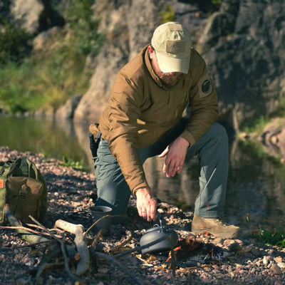 Mann kocht über einem Lagerfeuer an einem Flussufer und trägt eine Helikon-Tex® Wolfhound-Jacke – Climashield APEX 67 g.