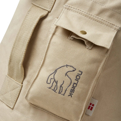 Nahaufnahme eines beigen Stoffes mit einem stilisierten Eisbärenlogo und dem Text „Nordisk®“ über einer Tasche, mit Knopfverschluss am Nordisk® Kongsberg 65, Duffle Rucksack.