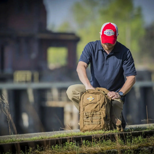 Ein Mann mit roter Mütze und blauem Hemd kniet neben einem Helikon-Tex® EDC-Rucksack – Cordura (21 Liter) auf Eisenbahnschienen mit Industriebauten im Hintergrund.