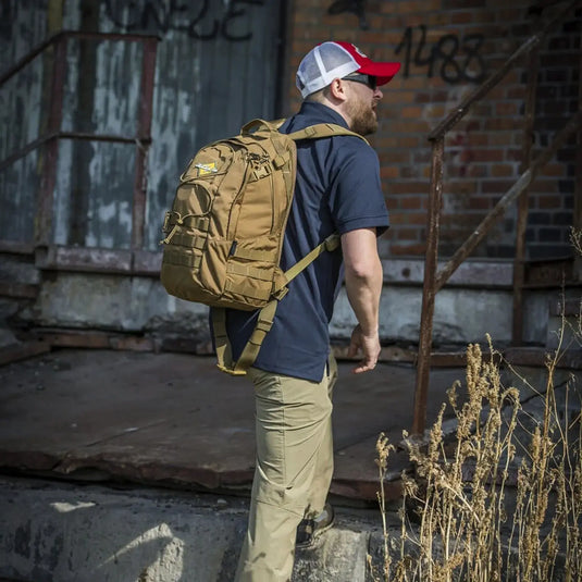 Mann mit einem Helikon-Tex® EDC-Rucksack – Cordura (21 Liter) geht die Treppe vor einem mit Graffiti markierten Gebäude hinauf.