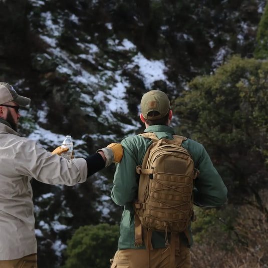 Zwei Personen auf einer Wanderung, wobei eine Person inmitten einer verschneiten Landschaft einer anderen Person eine Wasserflasche reicht, während sie robuste Helikon-Tex® RACCOON Mk2-Rucksäcke tragen, die für taktische Missionen entwickelt wurden.