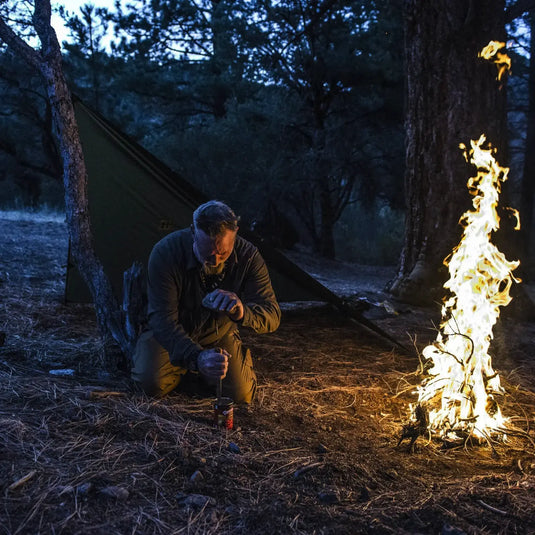 Eine Person, die in der Abenddämmerung an einem kleinen Campingkocher in der Nähe eines Lagerfeuers hockt, mit einem Helikon-Tex® SUPERTARP® Small-Zelt im Hintergrund.