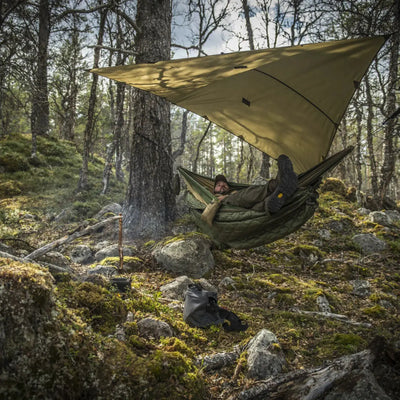 Eine Person ruht in einer Hängematte unter einem Helikon-Tex® SUPERTARP® Small, aufgestellt zwischen Bäumen in einem Wald, mit Campingausrüstung in der Nähe.