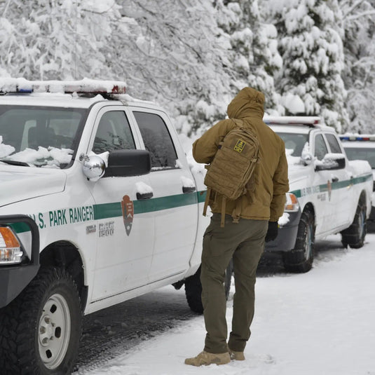 Eine Person in Winterkleidung und Helikon-Tex® Outdoor Tactical Pants – VERSASTRETCH®, die in einer verschneiten Umgebung in der Nähe von US-Park-Ranger-Fahrzeugen steht.