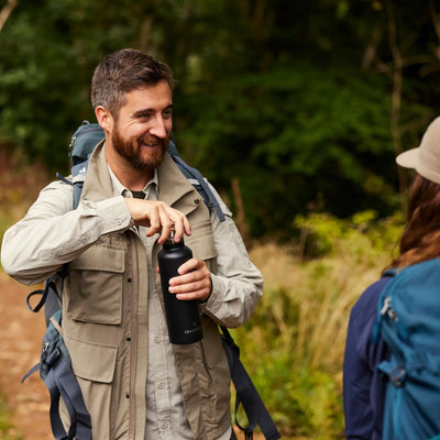 Ein Mann mit einem Rucksack von Craghoppers NosiLife Adventure Gilet IV für Herren, der lächelt und eine Wasserflasche in der Hand hält, während er draußen über Insektenschutzmöglichkeiten spricht.