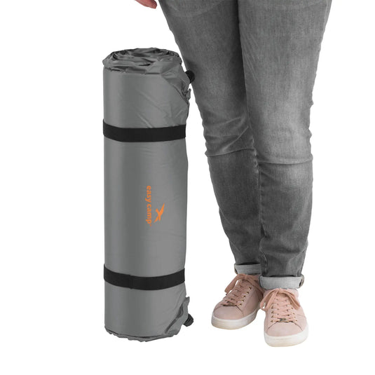 Stehende Person mit einer zusammengerollten Easy Camp® Siesta - Selbstaufblasende Isomatte in Grau neben ihren Beinen.