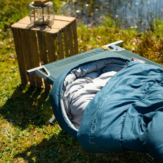 Eine Easy Camp® Pampas Camping-Bett auf einem leichten Gestell neben einem Holzstumpf mit einer Laterne draußen bei Tageslicht.