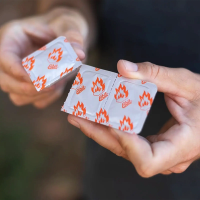 Hände halten eine Packung Kaugummi mit Flammenmuster auf der Umhüllung und Esbit® Brennstofftabletten 12x14g von Esbit®.