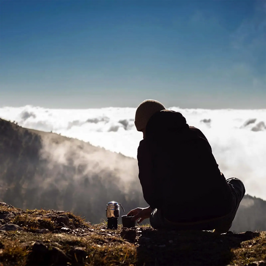 Person sitzt auf einem Berggipfel und genießt den Blick auf die Wolken darunter, bereit für ein Outdoor-Abenteuer mit Esbit® Brennstofftabletten 16x5g.