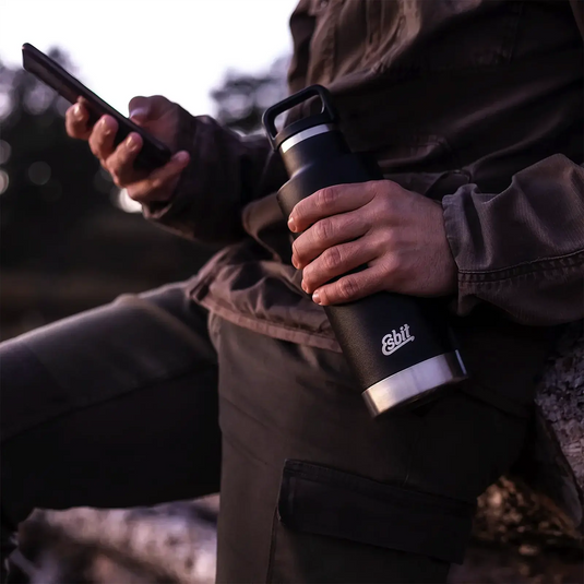 Person hält eine Esbit® Isolierflasche Sculptor „Standard Mouth“ 1000ml Edelstahl Schwarz in der Hand und nutzt ein Smartphone im Freien in der Abenddämmerung.