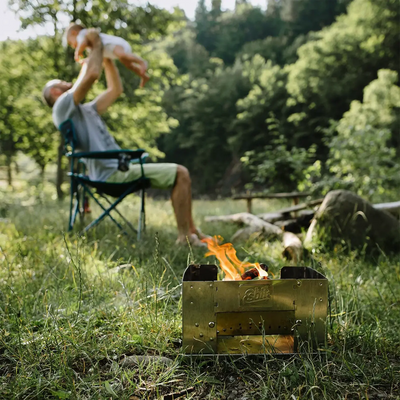 Eine Person sitzt in einem Campingstuhl und hebt ein Kleinkind über den Kopf, neben einem kleinen Lagerfeuer und einem klappbaren Esbit® Edelstahl Kohlegrill auf einer Rasenfläche.