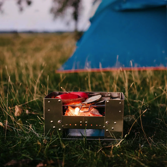 Tragbarer Esbit® Edelstahl Kohlegrill und Campingkocher mit einer Bratpfanne oben im Einsatz in der Nähe eines Zeltes im Freien in der Abenddämmerung.