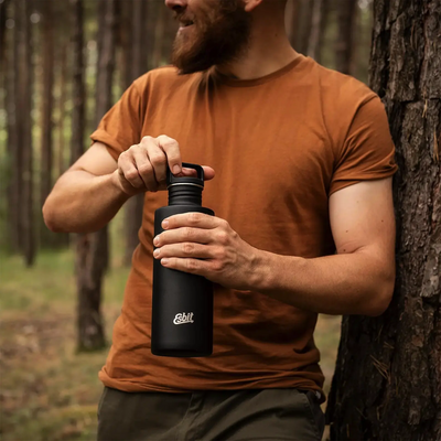 Ein bärtiger Mann in einem braunen T-Shirt öffnet im Wald eine schwarze Esbit® Trinkflasche Sculptor 1000 ml Edelstahl-Wasserflasche.