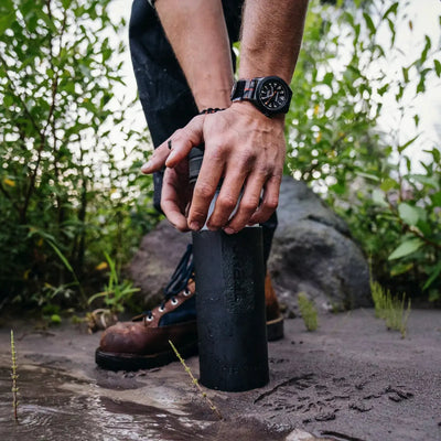 Person steht an einem Gewässer, hält einen Grayl Geopress Wasserfilter 710 ml fest, trägt eine Armbanduhr und braune Stiefel.