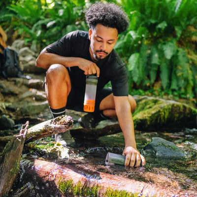 Mann sammelt Wasser aus einem Bach mit einem Grayl Ultrapress Wasserfilter 500 ml für eine effektive Wasserreinigung.