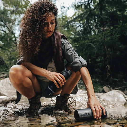 Satz mit Produktname und Markenname: Frau hockte an einem Bach und gefüllte einen Grayl® Ultrapress Wasserfilter 500ml für sauberes Trinkwasser.