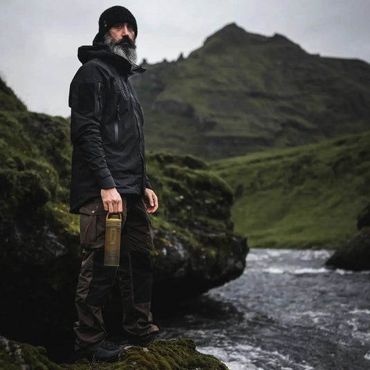 Bärtiger Mann steht an einem Bach in einer rauen Landschaft mit bewölktem Himmel und präsentiert den Grayl® Ultrapress Wasserfilter 500 ml für eine effektive Wasserreinigung.