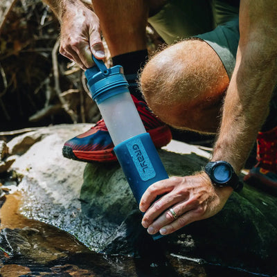 Mann füllt beim Wandern eine Wasserflasche aus einem Bach auf und verwendet einen Grayl® Ultrapress Wasserfilter 500 ml, um sauberes Trinkwasser zu gewährleisten.