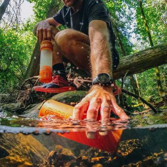 Mann reinigt Wasser aus einem Bach mit einem Grayl® Ultrapress Wasserfilter, während er mit dem Rucksack im Wald unterwegs ist.