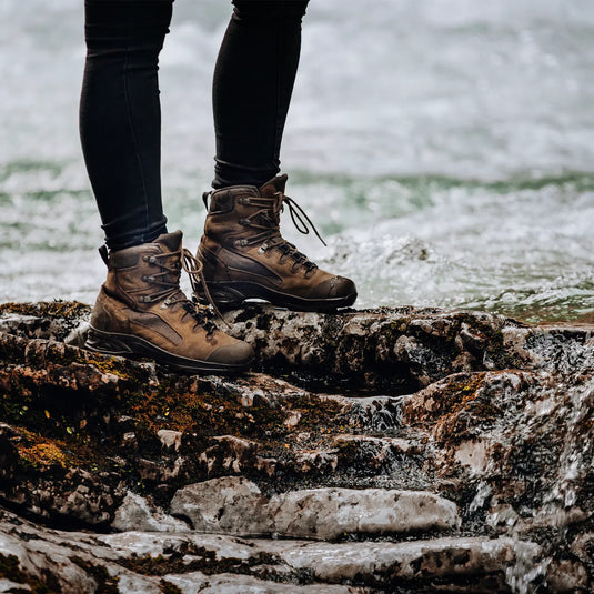 Person steht in HAIX® SCOUT 2 Wanderschuhen an einem felsigen Ufer, im Hintergrund Wasser.