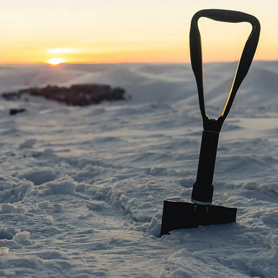 Ein HIGHLANDER® Doppel-Faltspaten, eingebettet im Schnee, mit einem Sonnenaufgang im Hintergrund.