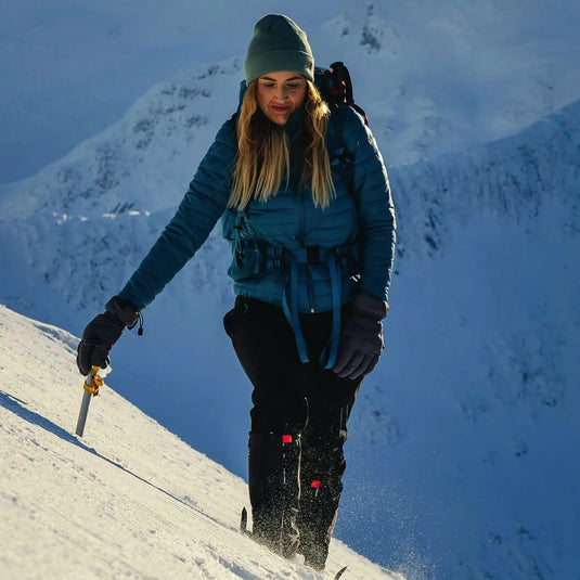 Ein Highlander erklimmt einen verschneiten Berghang, ausgerüstet mit einem Eispickel und HIGHLANDER® „Fara“ Isolationsjacke.