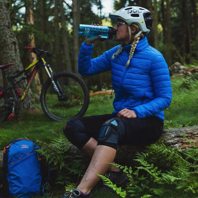 Ein Radfahrer macht eine Wasserpause im Wald, im Hintergrund seine HIGHLANDER® „Fara“ Isolationsjacke.