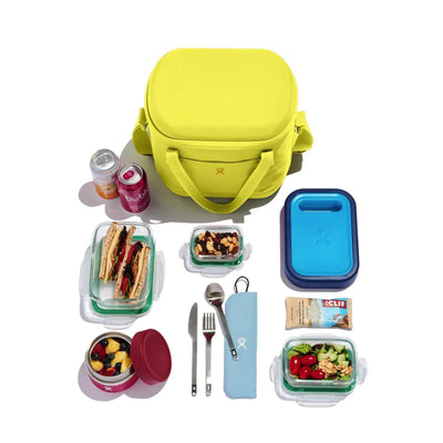 Verschiedene Mittagsgerichte, darunter ein Sandwich, Salat, Snacks und ein Hydro Flask® Carry Out™ Soft Cooler, ordentlich angeordnet mit Utensilien und Behältern.