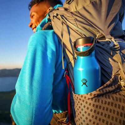 Wanderer mit einer blauen Hydro Flask® 21 oz (621 ml) Lightweight Standard Mouth Trail Series™ an einem Rucksack befestigt, genießt die Aussicht bei Sonnenuntergang.