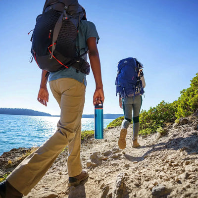 Zwei Wanderer mit Rucksäcken und Hydro Flask® 24 oz (710 ml) Lightweight Wide Mouth Trail Series™ wandern entlang eines Küstenpfades.