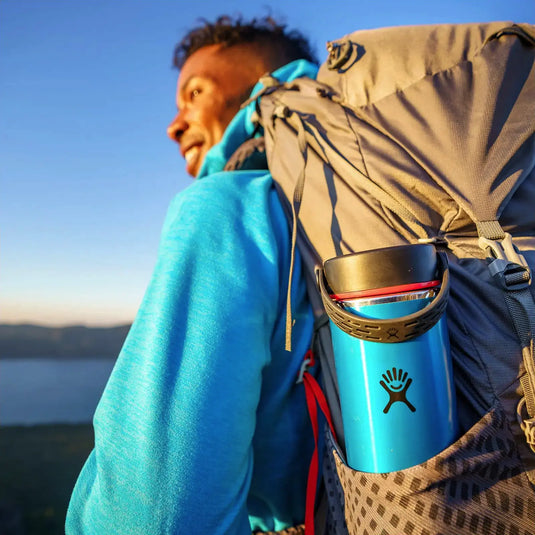 Wanderer mit Rucksack und einer Hydro Flask® 24 oz (710 ml) Lightweight Wide Mouth Trail Series™ genießt eine malerische Aussicht bei Sonnenuntergang.