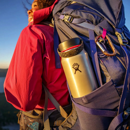 Eine Nahaufnahme eines leichten Wanderrucksacks mit einer Hydro Flask® 40 oz (1183 ml) Lightweight Wide Mouth Trail Series™ und daran befestigten Karabinern in der Abenddämmerung.